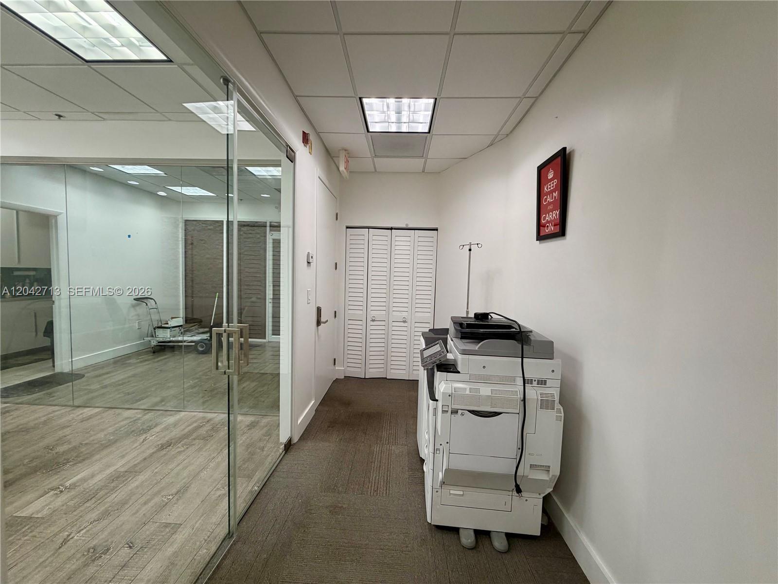 A hallway features a glass-walled office, a large printer, and a wall-mounted sign — 7500 NW 25th St # 205, Miami FL 33122, MIAMI, FL