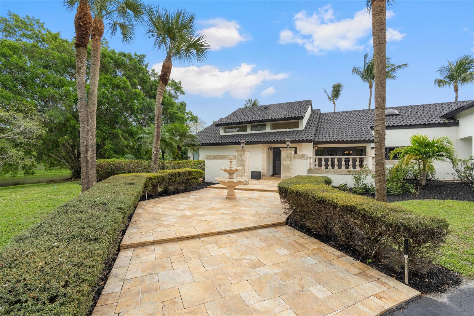 A stone-paved walkway with a central fountain leads to the entrance of a white house — 10076 El Caballo Court, Delray Beach FL 33446, DELRAY BEACH, FL