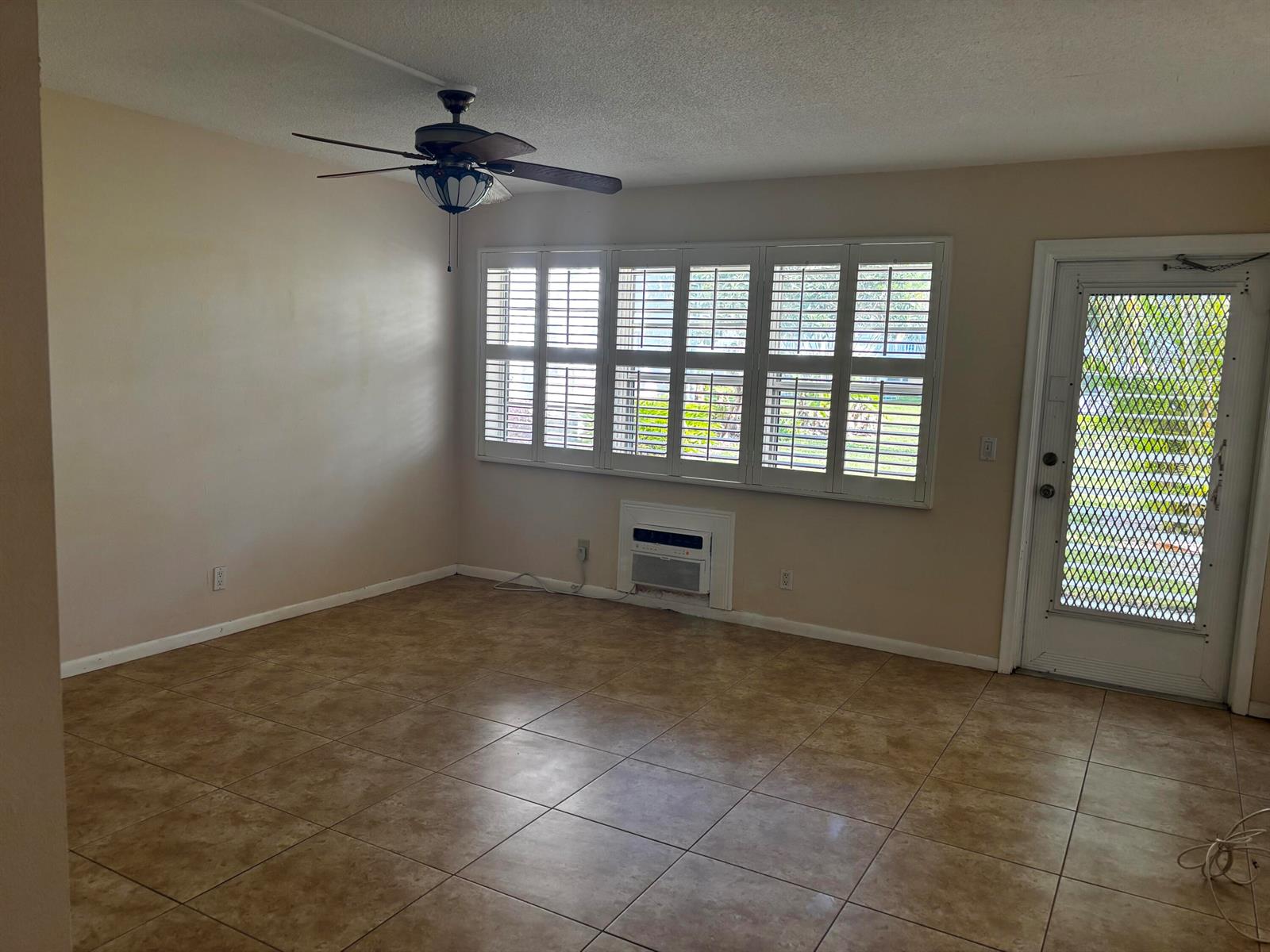 An empty room features tan tile flooring, a ceiling fan, plantation shutters, and an exterior door — 284 Windsor M, West Palm Beach FL 33417, WEST PALM BEACH, FL