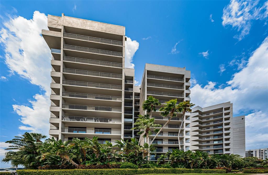 A low-angle view of a multi-story concrete apartment building with balconies surrounded by tropical landscaping — 1660 GULF BOULEVARD UNIT 208, CLEARWATER BEACH, FL