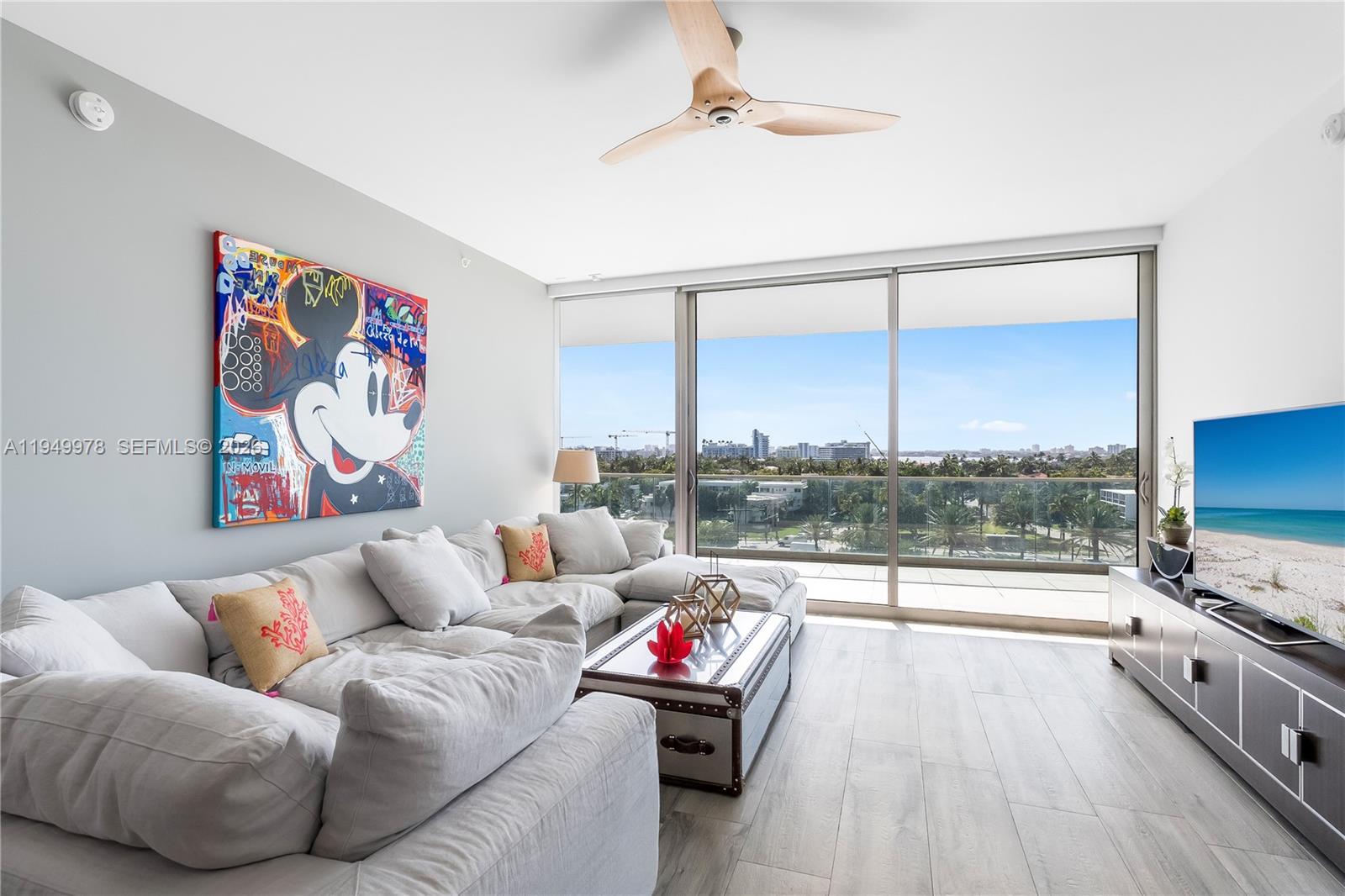 A living room features a large sectional sofa, a Mickey Mouse painting, and floor-to-ceiling windows — 10201 Collins Ave # 605, Bal Harbour FL 33154, BAL HARBOUR, FL