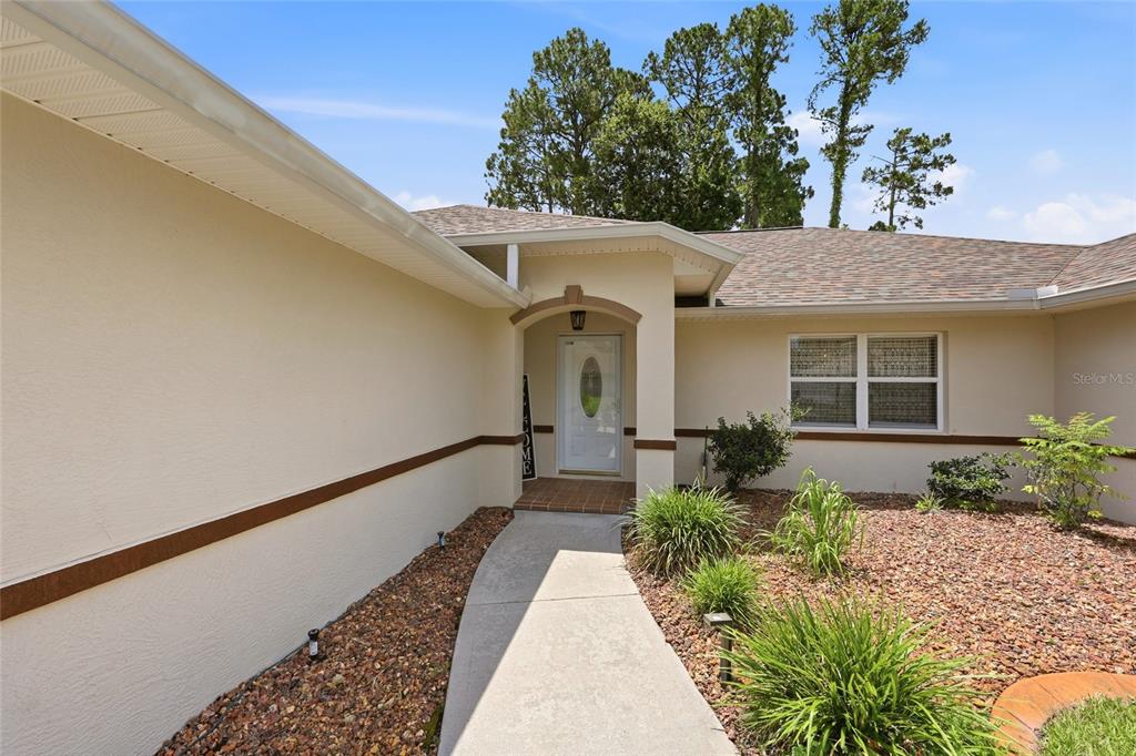 A concrete walkway leads to the front entrance of a beige house with brown trim — 56 BASSETT LANE, PALM COAST, FL