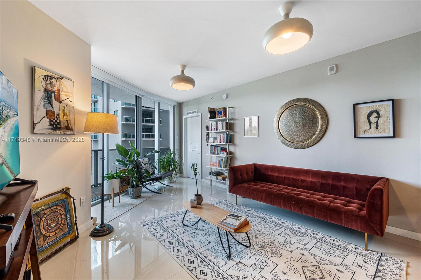 A modern living room features a velvet sofa, patterned rug, bookshelf, and floor-to-ceiling windows — 488 NE 18th St # 401, Miami FL 33132, MIAMI, FL