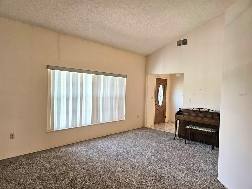 A living room with gray carpet, a large window with vertical blinds, and an upright piano — 1850 CHAUCER WAY, KISSIMMEE, FL