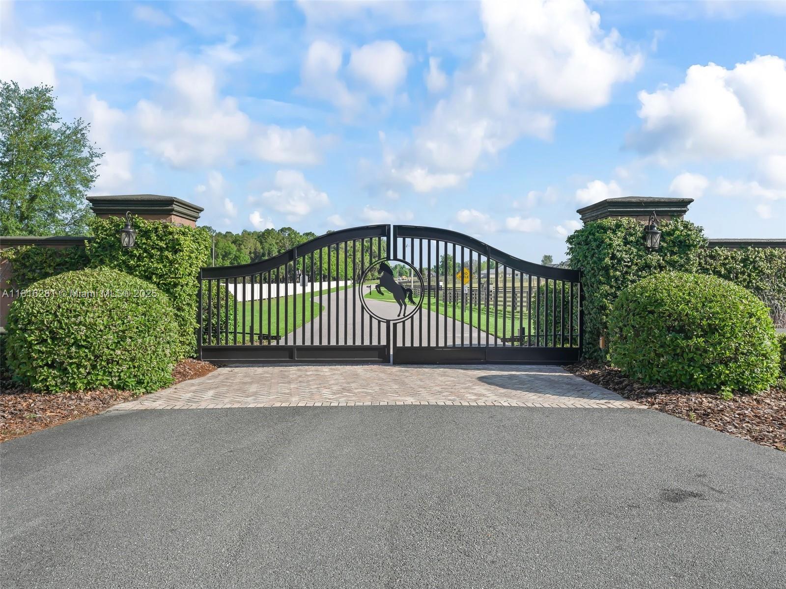 A black metal gate with a horse emblem stands between two stone pillars and hedges — 7161 NE 22nd Ct Rd, Ocala FL 34479, OCALA, FL
