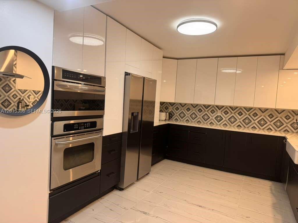 A modern kitchen features white upper cabinets, dark lower cabinets, stainless steel appliances, and patterned backsplash — 500 Three Islands Blvd # 106, Hallandale Beach FL 33009, HALLANDALE BEACH, FL