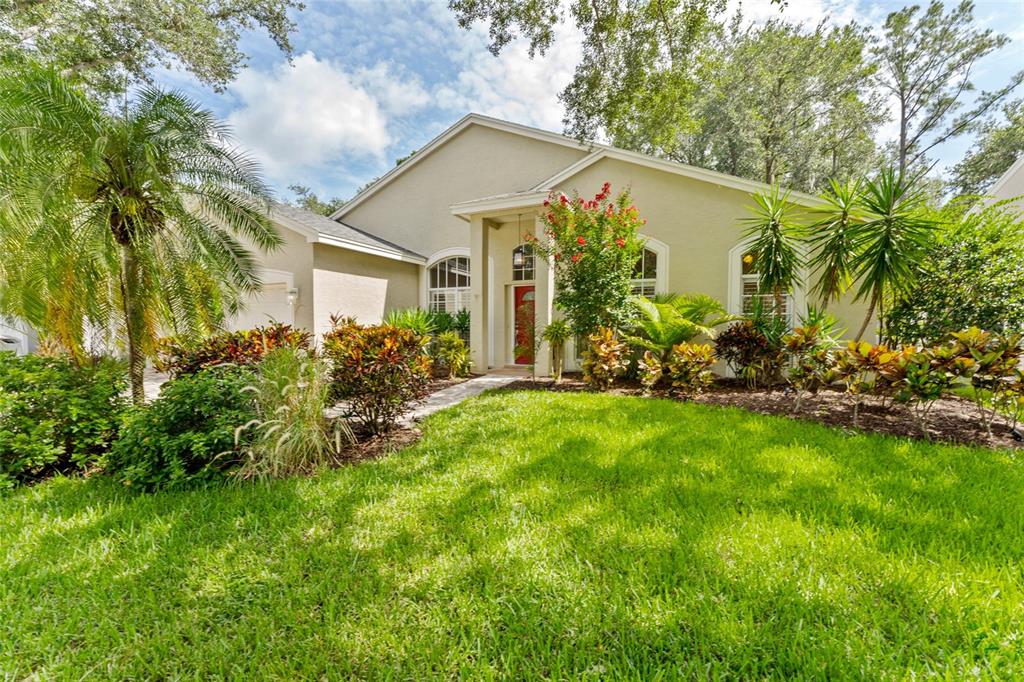 A single-story house with a red front door and lush landscaping under a blue sky — 4287 TREMBLAY WAY, PALM HARBOR, FL