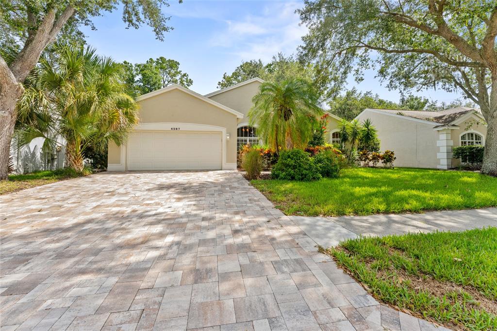 A beige single-story house features a paved driveway, a two-car garage, and lush landscaping — 4287 TREMBLAY WAY, PALM HARBOR, FL