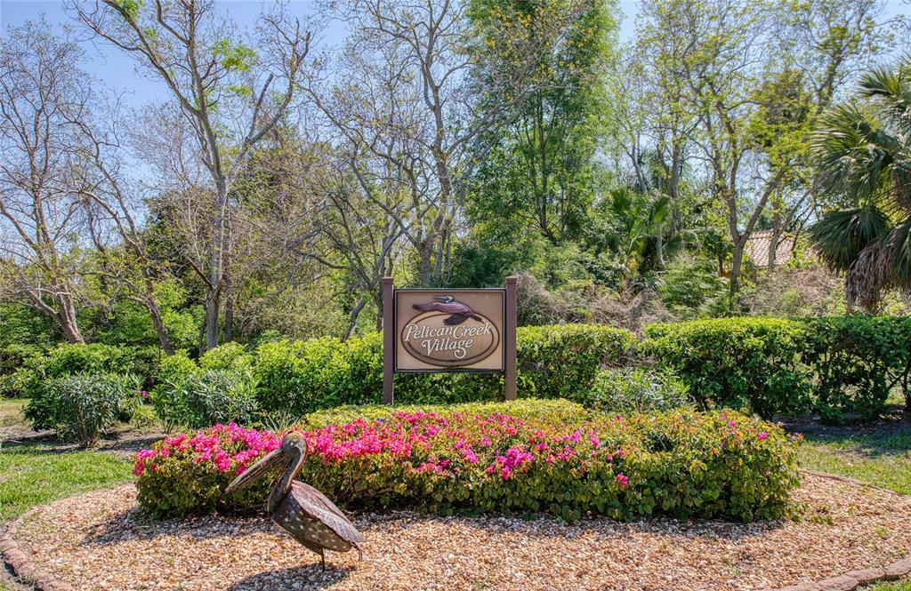 A sign for Pelican Creek Village stands in a garden bed with a pelican statue — 6305 PELICAN CREEK CROSSING UNIT D, ST PETERSBURG, FL