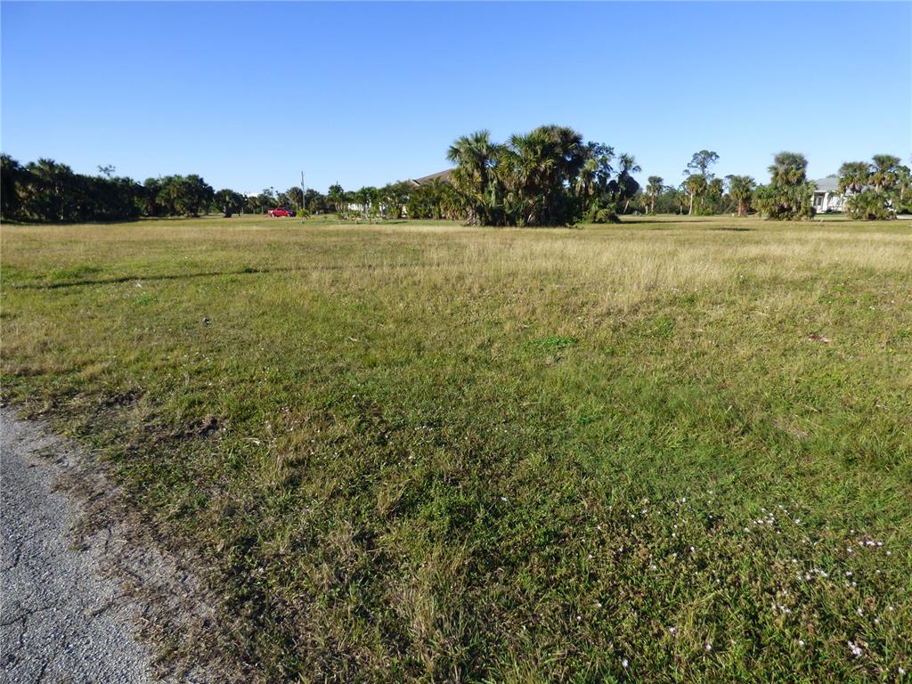 A wide, grassy vacant lot under a clear blue sky with distant trees and houses — 24378 GIJON LANE, PUNTA GORDA, FL