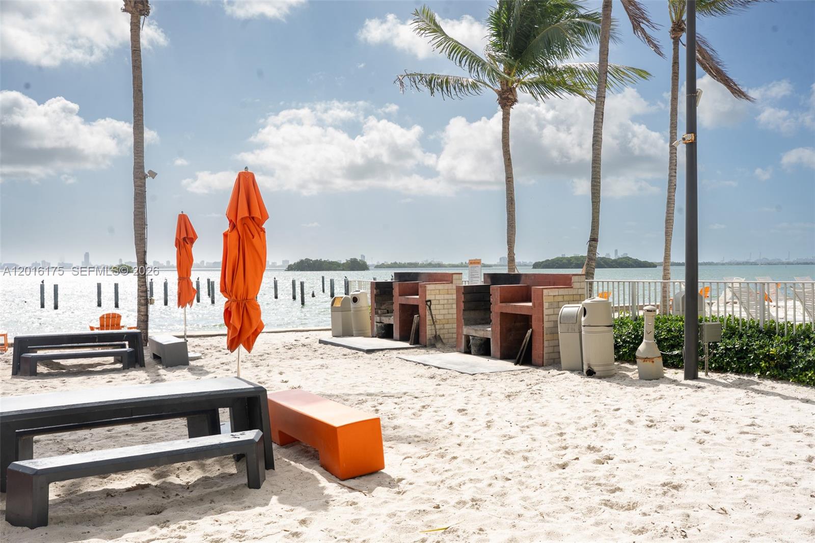 A sandy beach area features picnic tables, orange umbrellas, and brick barbecue grills near water — 750 NE 64th St # B215, Miami FL 33138, MIAMI, FL