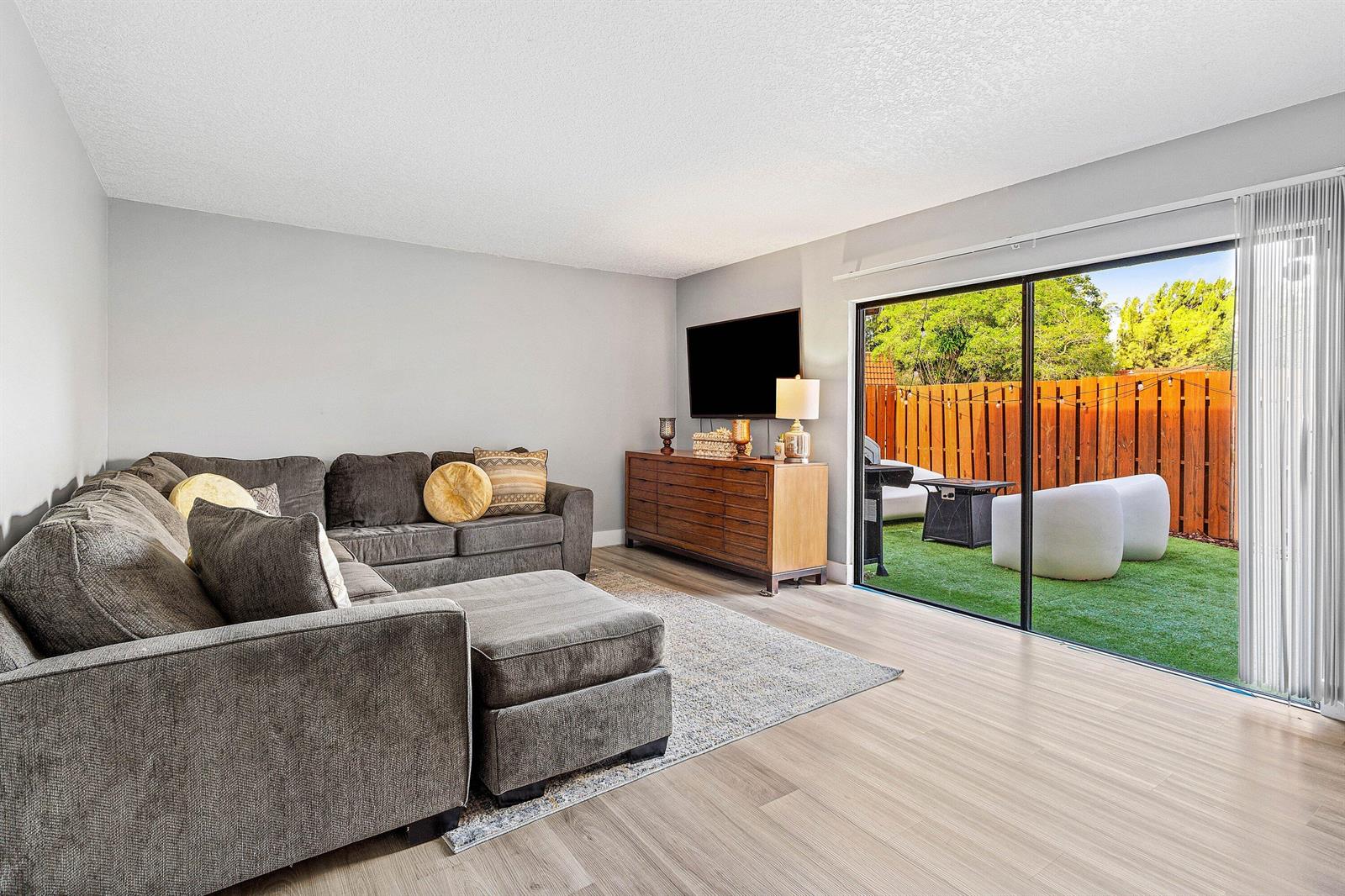 A living room with a gray sectional sofa, wood media console, and sliding glass patio doors — 3425 San Bernadino Drive # b, Delray Beach FL 33445, DELRAY BEACH, FL