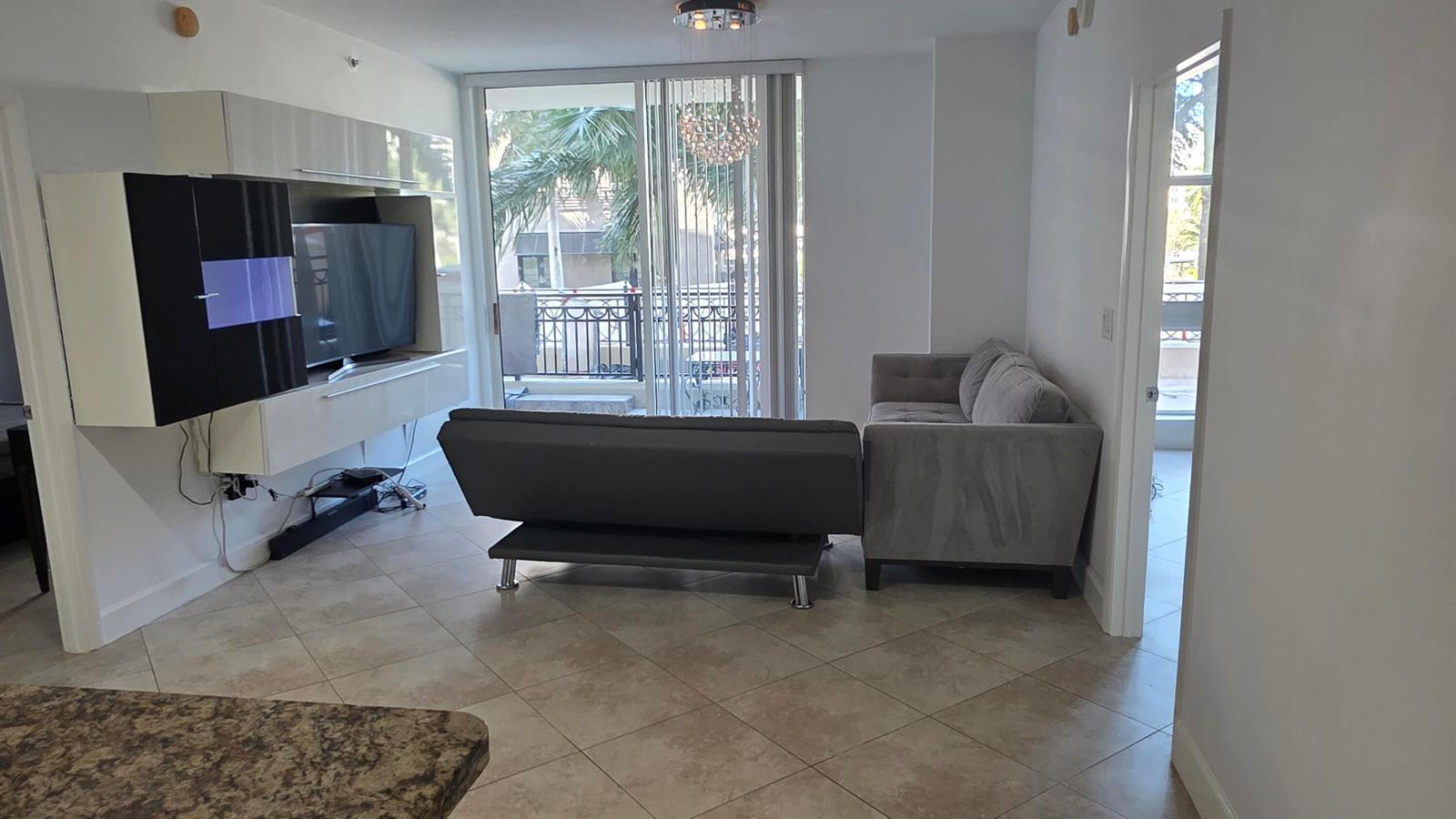 A living room features a gray sofa, a television on a wall unit, and balcony access — 550 Okeechobee Boulevard # 308, West Palm Beach FL 33401, WEST PALM BEACH, FL