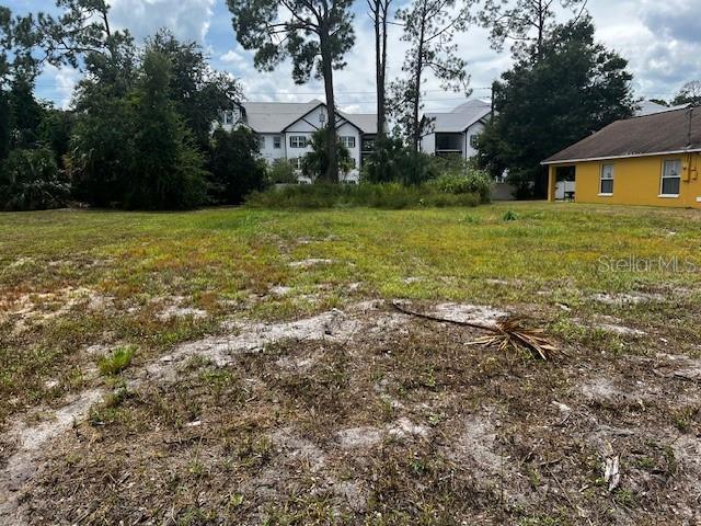 A vacant, grassy lot with sandy patches sits in front of trees and nearby buildings — IOWA STREET, DAYTONA BEACH, FL