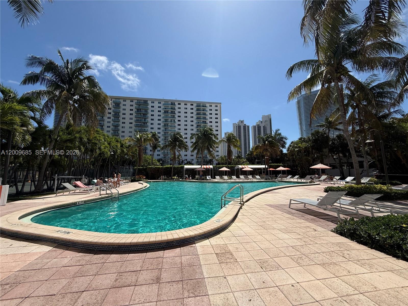 A large outdoor swimming pool is surrounded by lounge chairs, palm trees, and tall buildings — SUNNY ISLES BEACH, FL