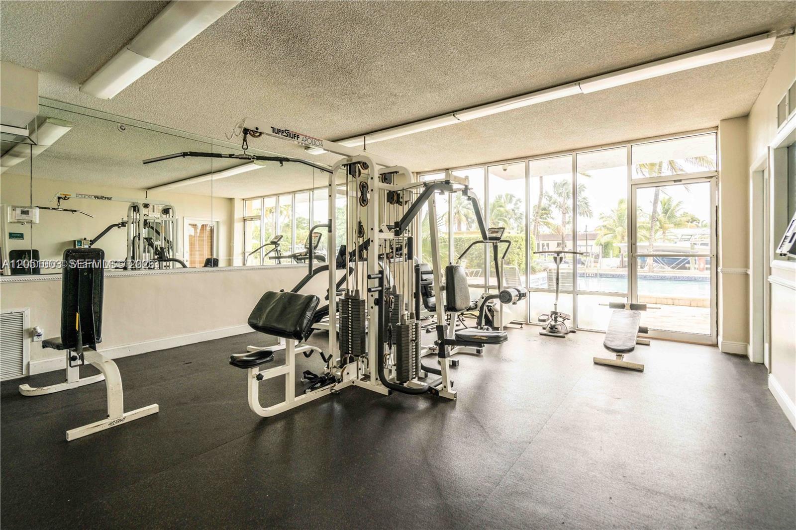 A gym room features a multi-station weight machine, rubber flooring, mirrored walls, and floor-to-ceiling windows — 2350 NE 135th St # 403, North Miami FL 33181, NORTH MIAMI, FL