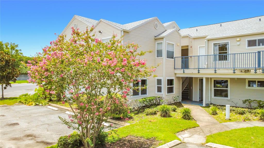 A two-story beige apartment building with a flowering tree in the foreground and parking area — 2504 WOODGATE BOULEVARD UNIT 203, ORLANDO, FL