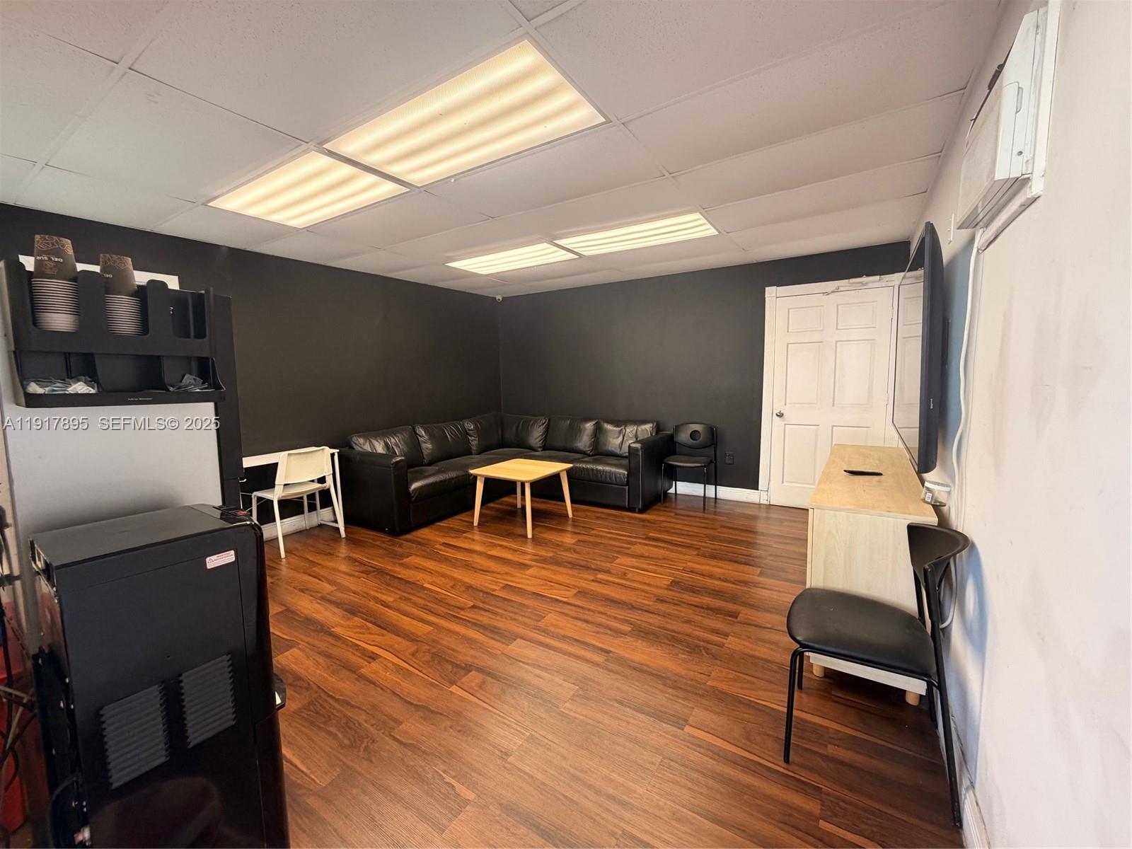 A room with dark walls, a black sectional sofa, wood flooring, and fluorescent ceiling lights — 7298 NW 54th St, Miami FL 33166, MIAMI, FL