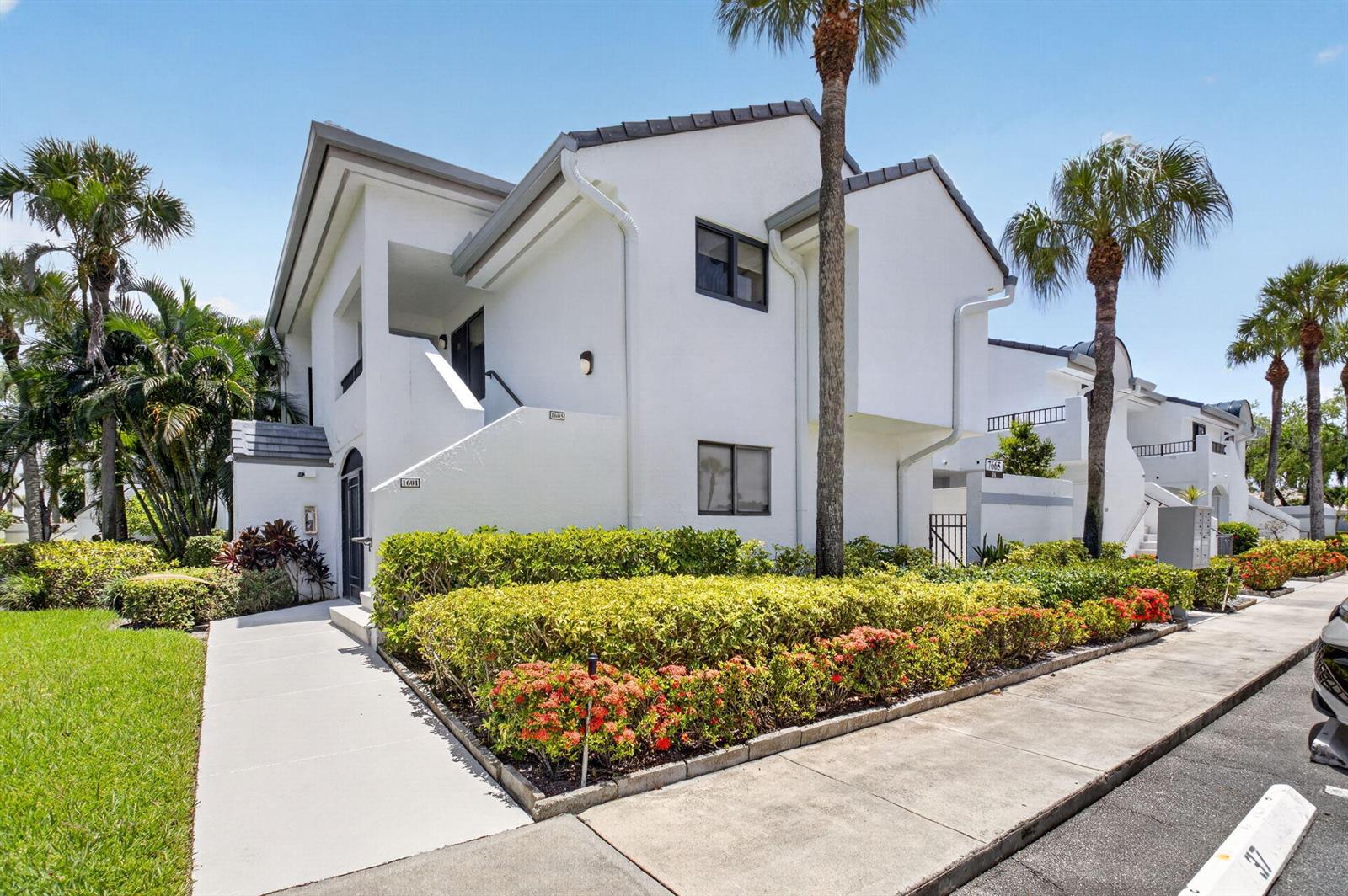 A two-story white stucco building with a walkway, landscaping, and palm trees under a blue sky — 7665 Glendevon Lane # 1605, Delray Beach FL 33446, DELRAY BEACH, FL