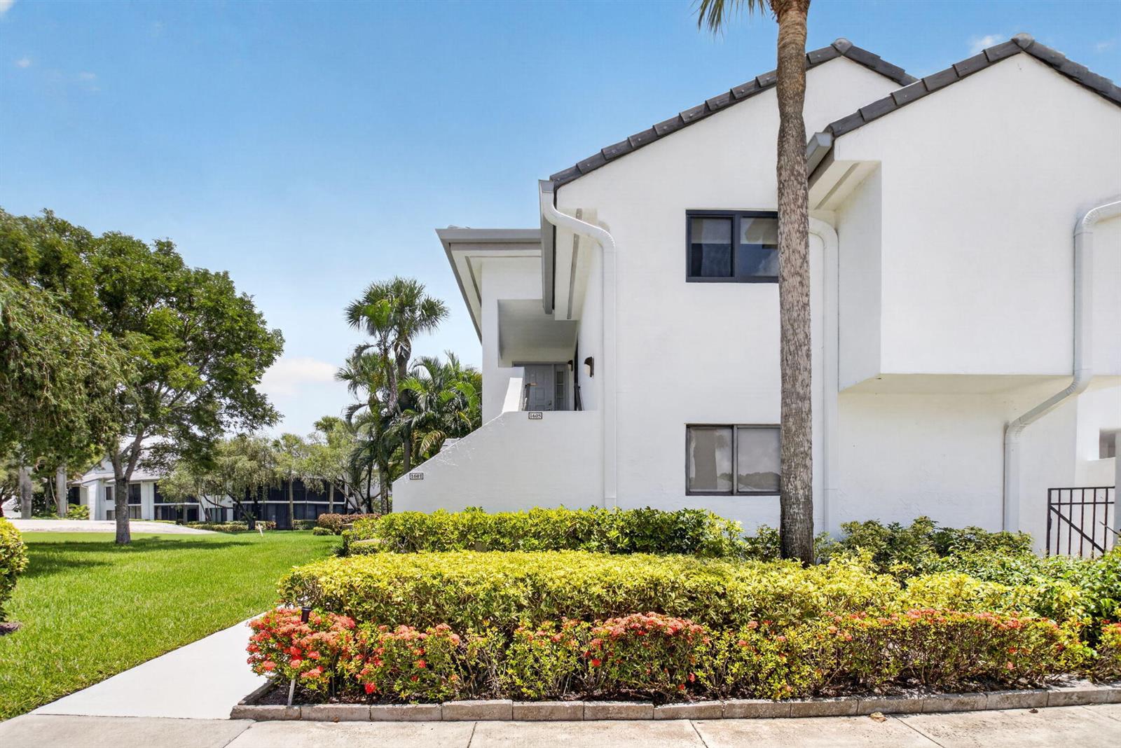 A white two-story residential building features a landscaped garden with red flowers in the foreground — 7665 Glendevon Lane # 1605, Delray Beach FL 33446, DELRAY BEACH, FL