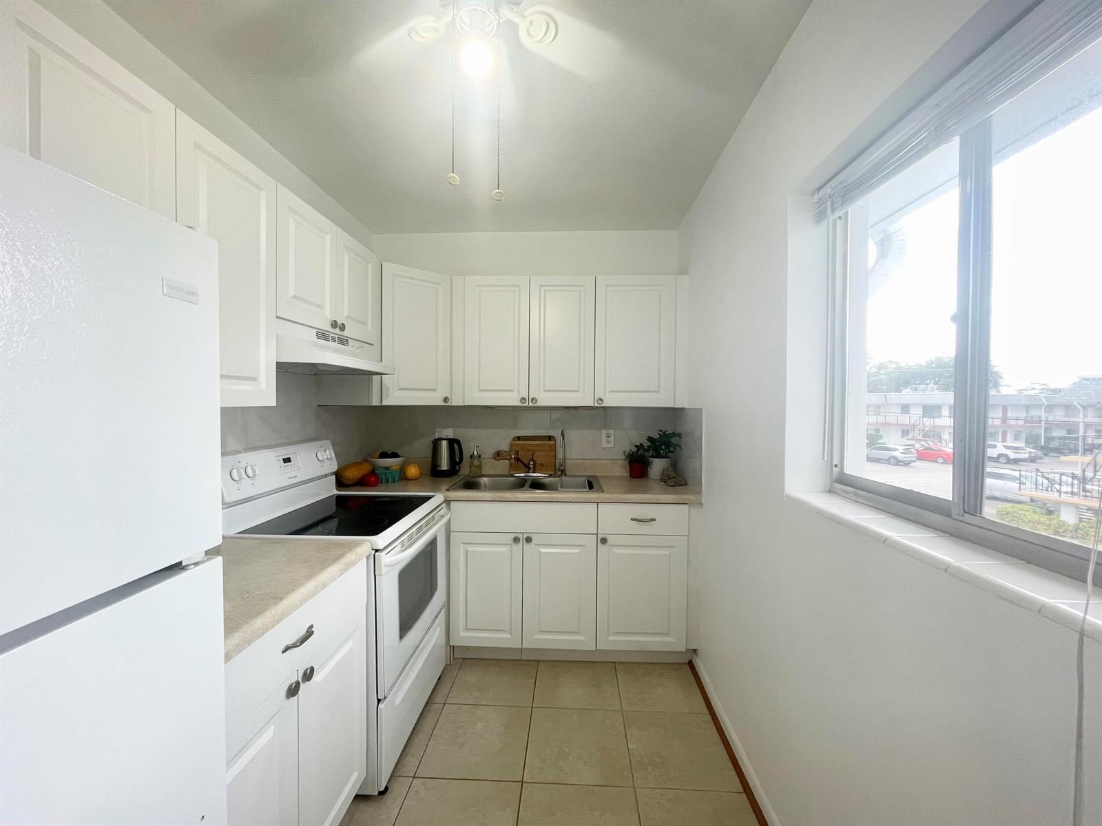 A galley kitchen features white cabinets, white appliances, beige tile flooring, and a large window — 2920 Cynthia Lane # 203, FL 33461, FL