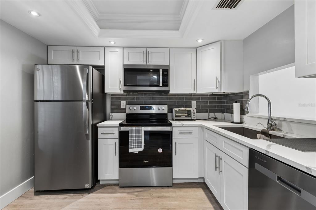 A kitchen features white cabinets, stainless steel appliances, a gray tile backsplash, and wood flooring — 2460 NORTHSIDE DRIVE UNIT 604, CLEARWATER, FL