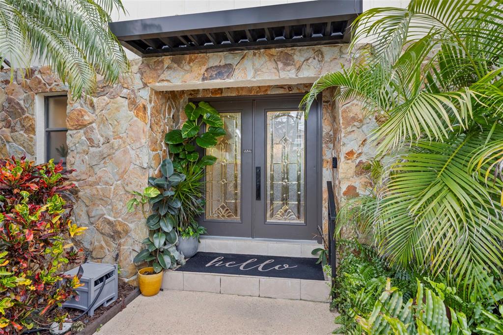 A stone-clad entryway features double glass-paneled doors, a black awning, and lush tropical landscaping — 93 S CANAL DRIVE, PALM HARBOR, FL