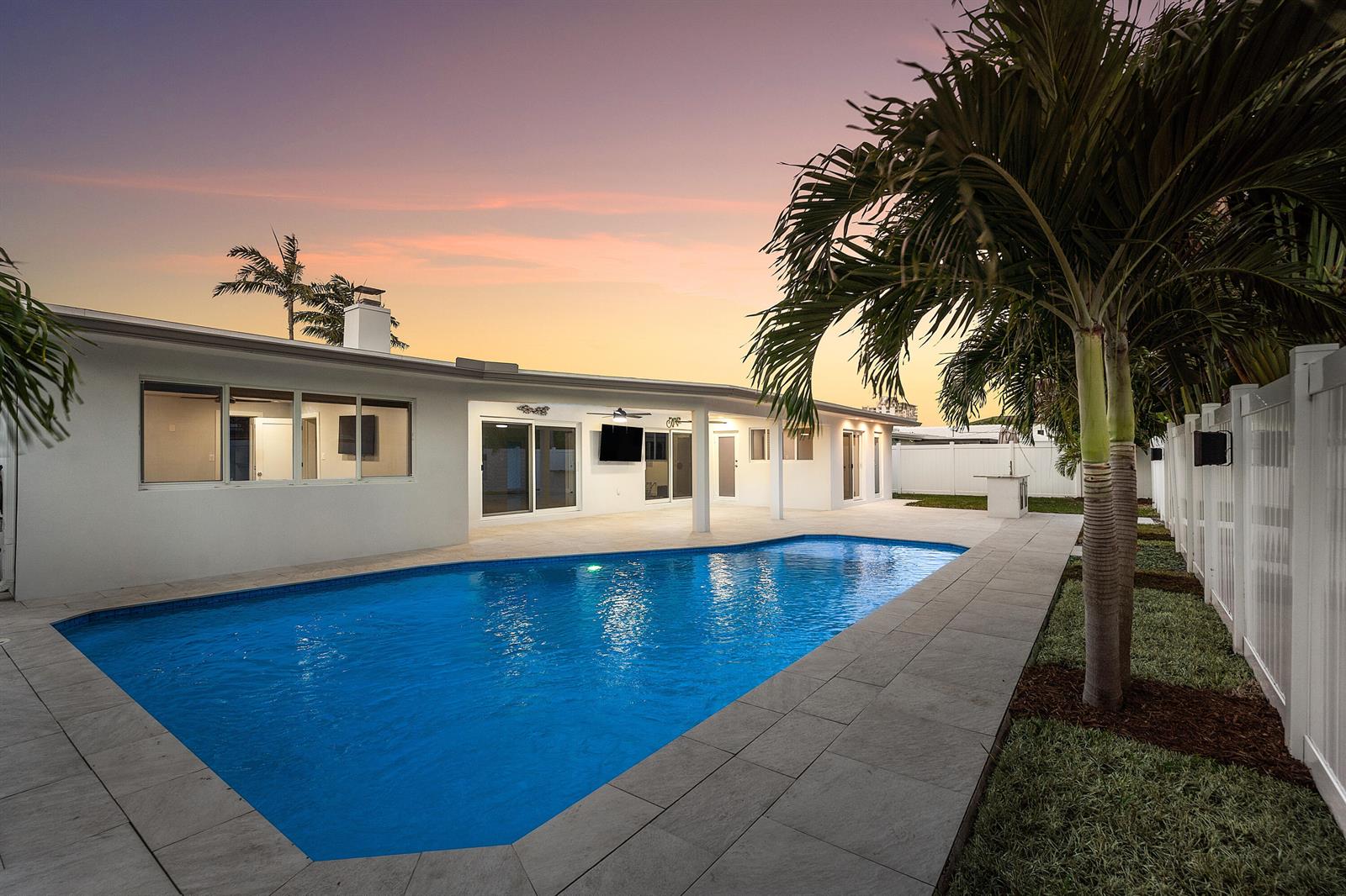 A backyard swimming pool with a stone patio sits behind a white house at sunset — 2846 NE 35th Court, Lighthouse Point FL 33064, LIGHTHOUSE POINT, FL