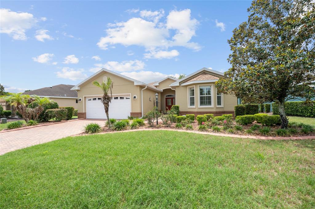 A single-story tan house with a two-car garage, brick driveway, and a large front tree — 9274 SW 94TH LOOP, OCALA, FL
