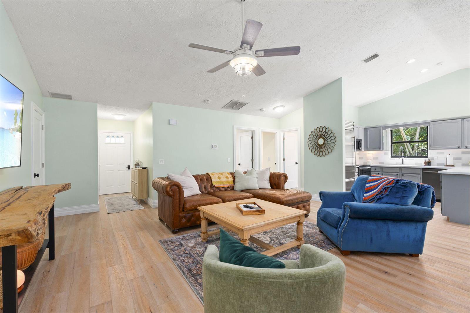 A living room features a brown leather sectional, blue armchair, light wood floors, and kitchen — 1904 NE Avenida Dracaena, Jensen Beach FL 34957, JENSEN BEACH, FL