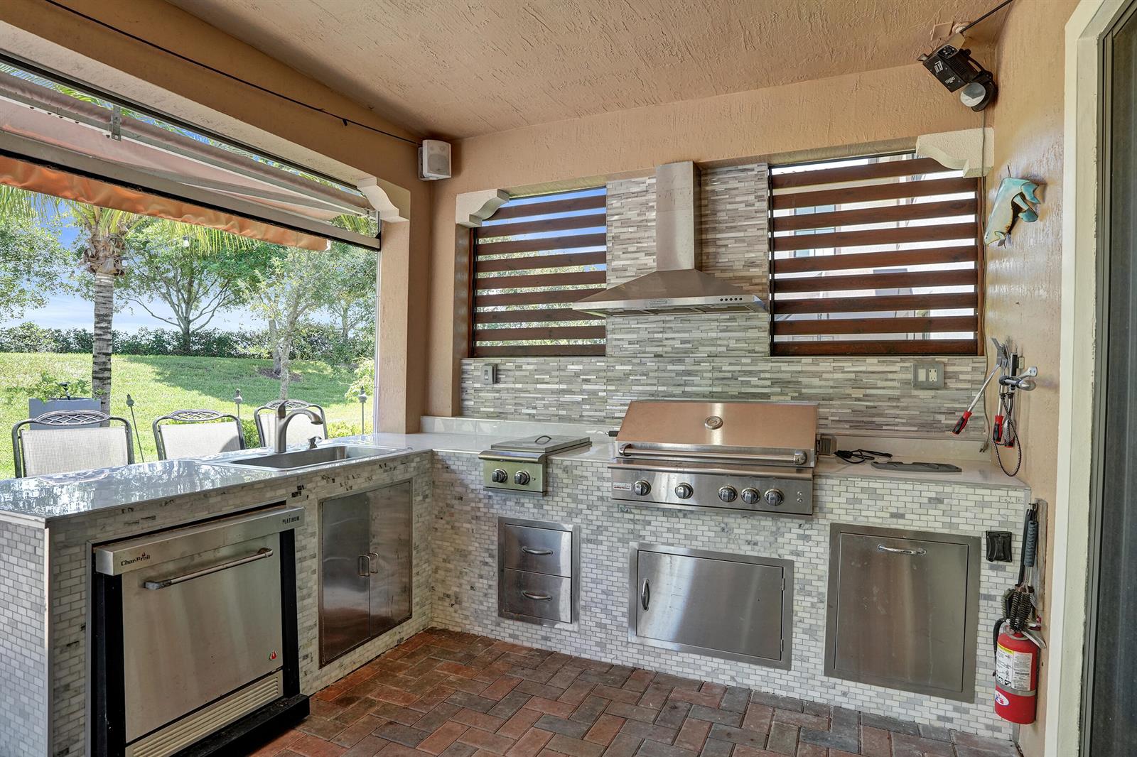 An outdoor kitchen features a stainless steel grill, sink, stone tile backsplash, and brick flooring — 11405 Solstice Circle, Parkland FL 33076, PARKLAND, FL