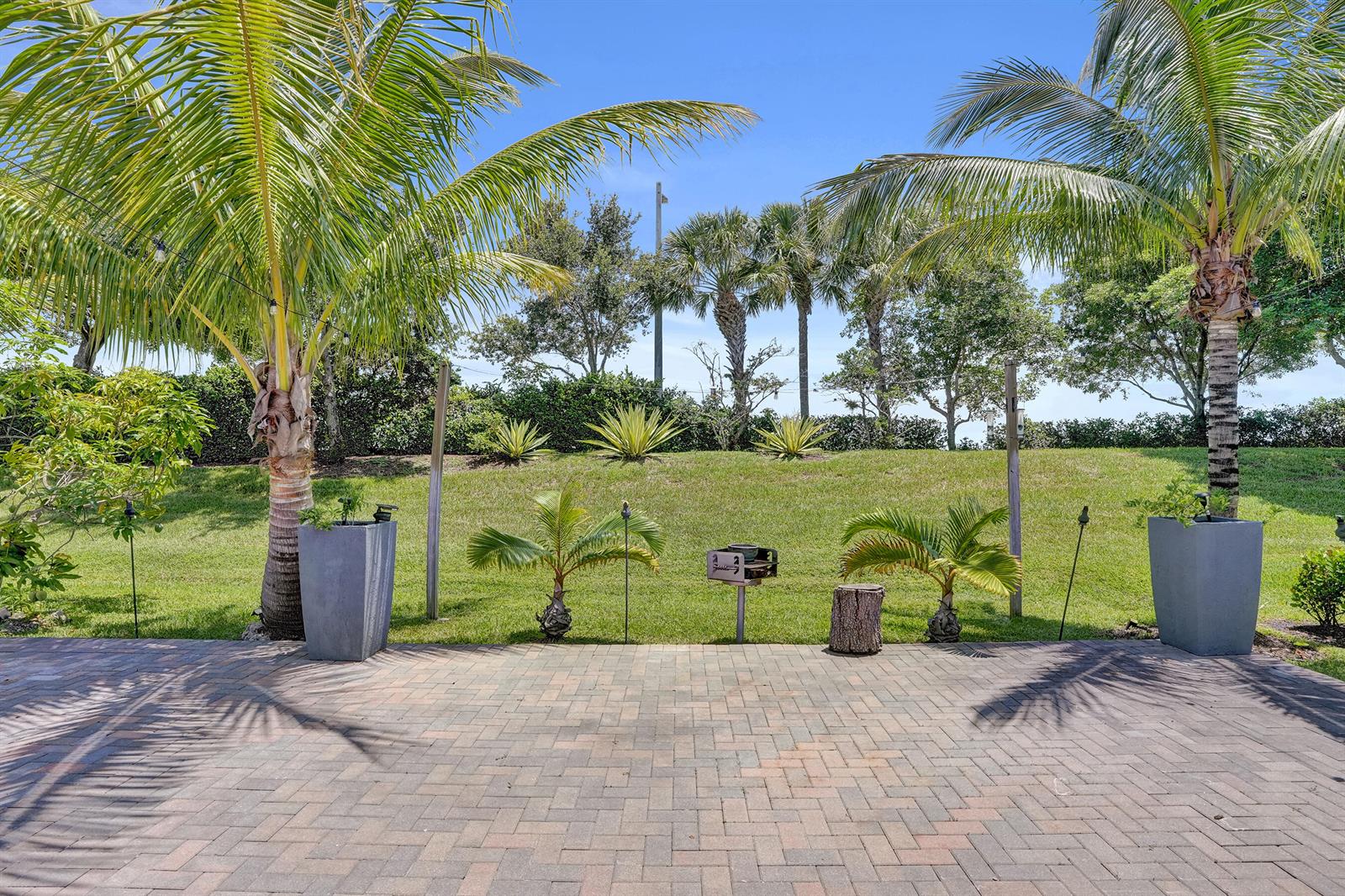 A paved patio overlooks a grassy backyard with palm trees and a small charcoal grill — 11405 Solstice Circle, Parkland FL 33076, PARKLAND, FL