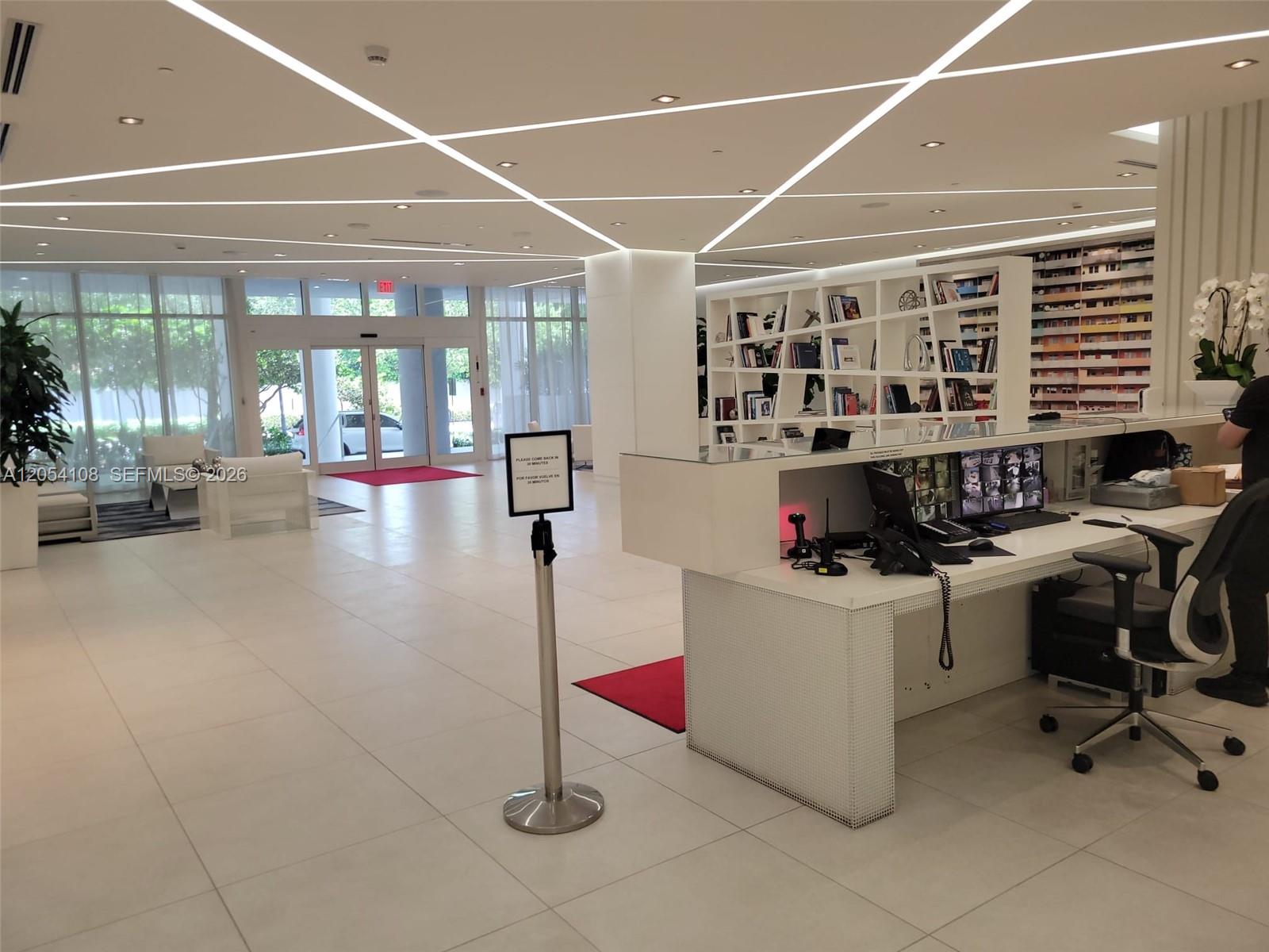 A modern lobby features a white reception desk, geometric ceiling lighting, and a bookshelf partition — 4250 Biscayne Blvd # 806, Miami FL 33137, MIAMI, FL