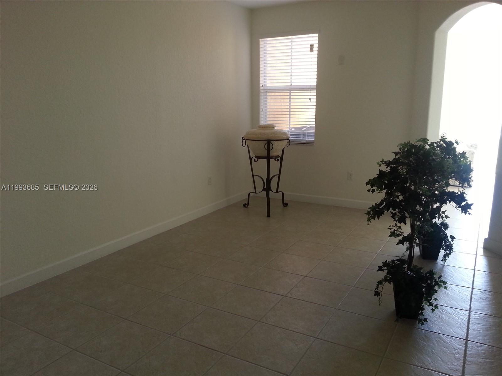 An empty room features beige tile flooring, a window with blinds, and two potted plants — 11050 NW 86th Ter, Doral FL 33178, DORAL, FL