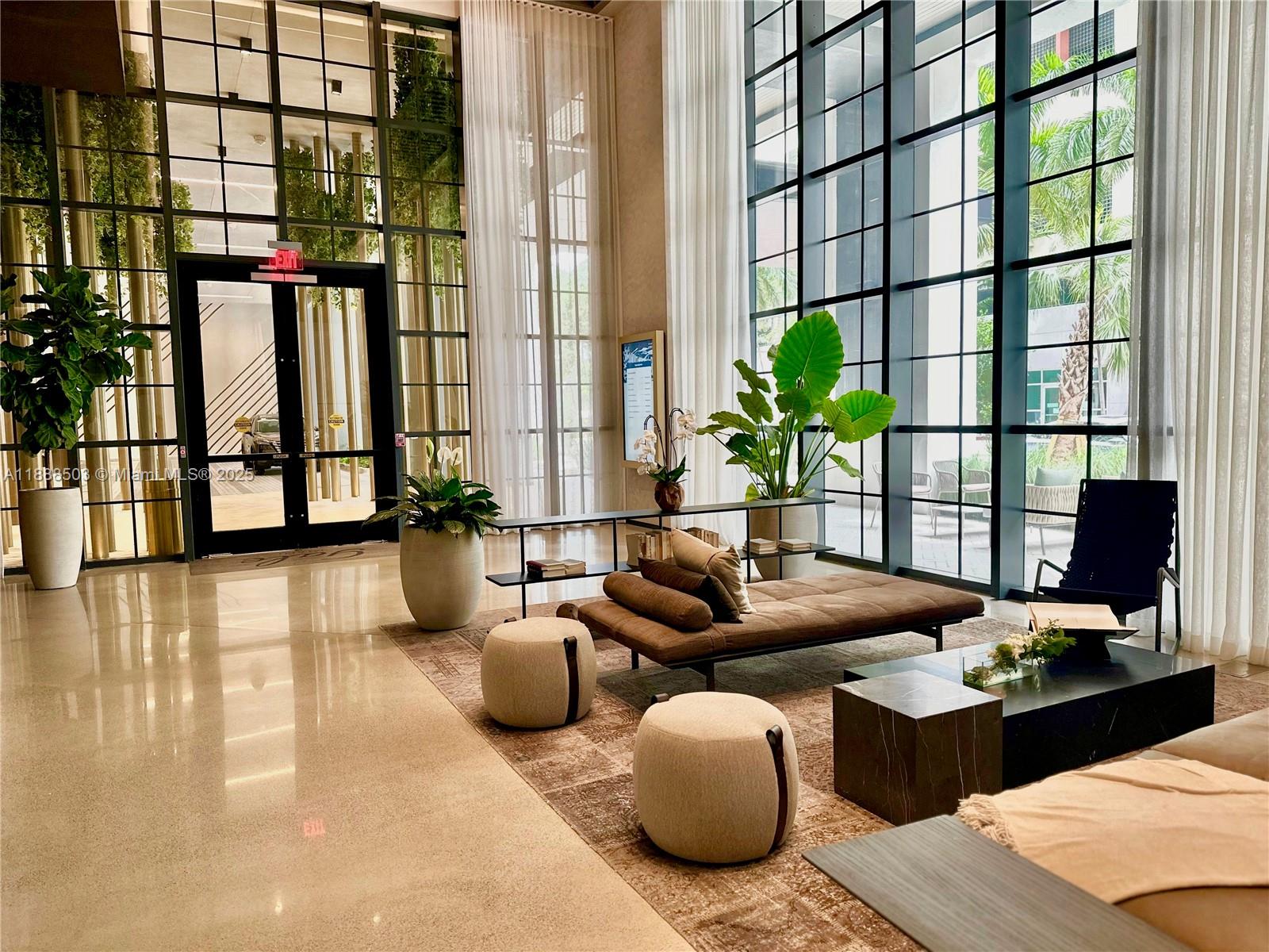 A modern lobby features floor-to-ceiling windows, a brown daybed, neutral ottomans, and potted plants — 159 NE 6th St # 3306, Miami FL 33132, MIAMI, FL