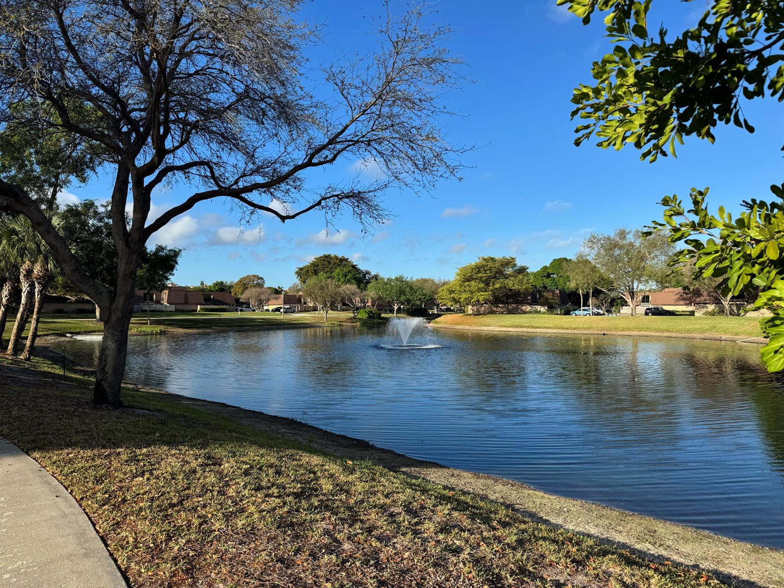 A pond with a central fountain is surrounded by trees and residential buildings under blue skies — 219 2nd Lane, Palm Beach Gardens FL 33418, PALM BEACH GARDENS, FL