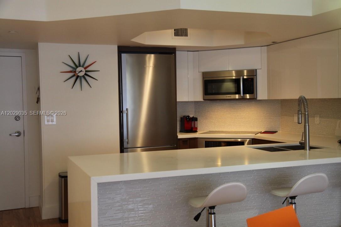 A modern kitchen features white cabinetry, stainless steel appliances, and a breakfast bar with stools — 8911 Collins Ave # 1104, Surfside FL 33154, SURFSIDE, FL