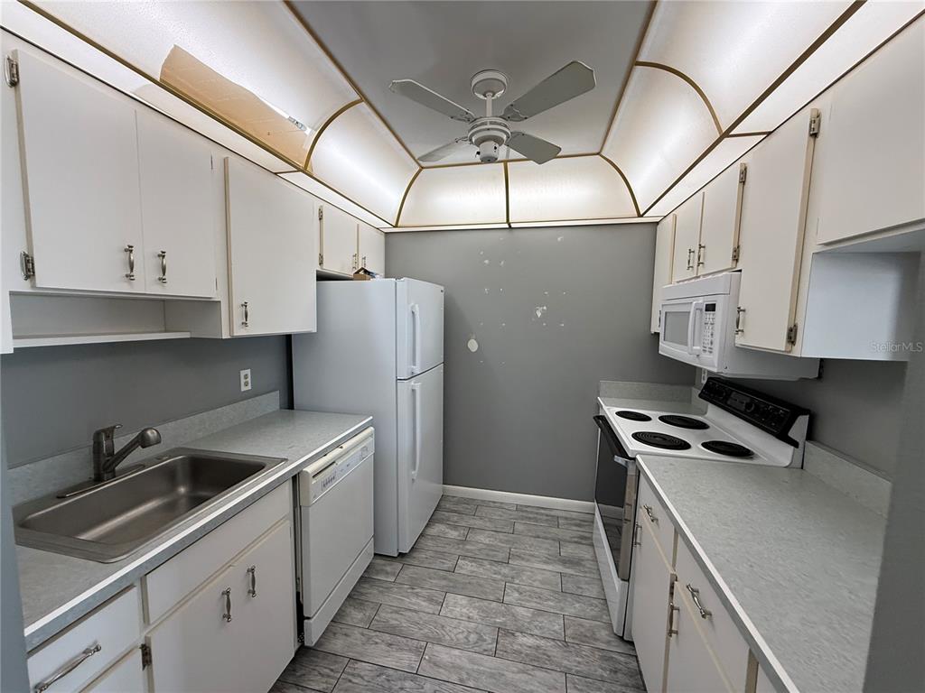 A galley kitchen features white cabinets, white appliances, gray walls, and wood-look tile flooring — 22 BIRDIE DRIVE, NEW SMYRNA BEACH, FL