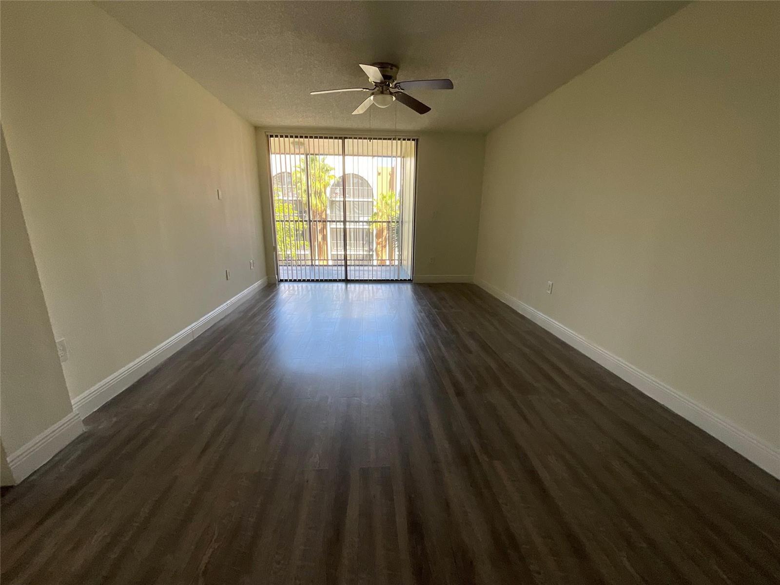 An empty room features dark wood-look flooring, beige walls, a ceiling fan, and sliding glass doors — 18335 NW 68 Avenue # 516, Hialeah FL 33015, HIALEAH, FL