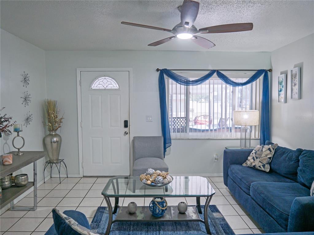 A living room features a blue sofa, glass coffee table, tile flooring, and a ceiling fan — 6436 CARDINAL DR, CLEARWATER, FL