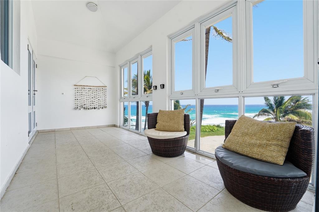 A sunroom with tile floors and two wicker chairs overlooks the ocean through large windows — MARFIL ST. TESORO DEL MAR IN LAS PALMAS, HATILLO, PR