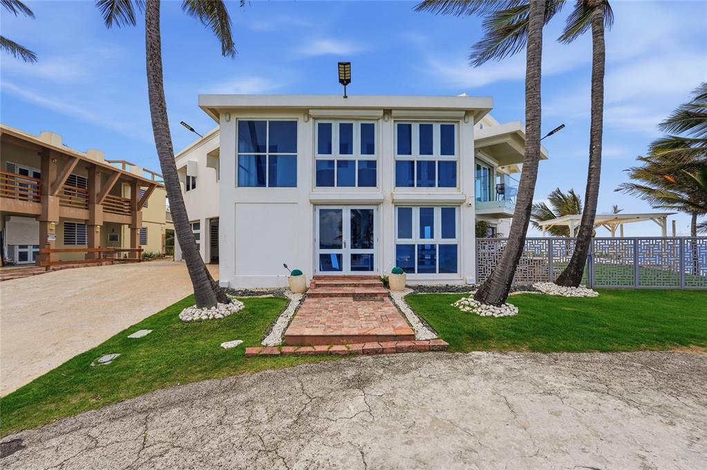 A two-story white house with large windows and a brick walkway sits between palm trees — MARFIL ST. TESORO DEL MAR IN LAS PALMAS, HATILLO, PR
