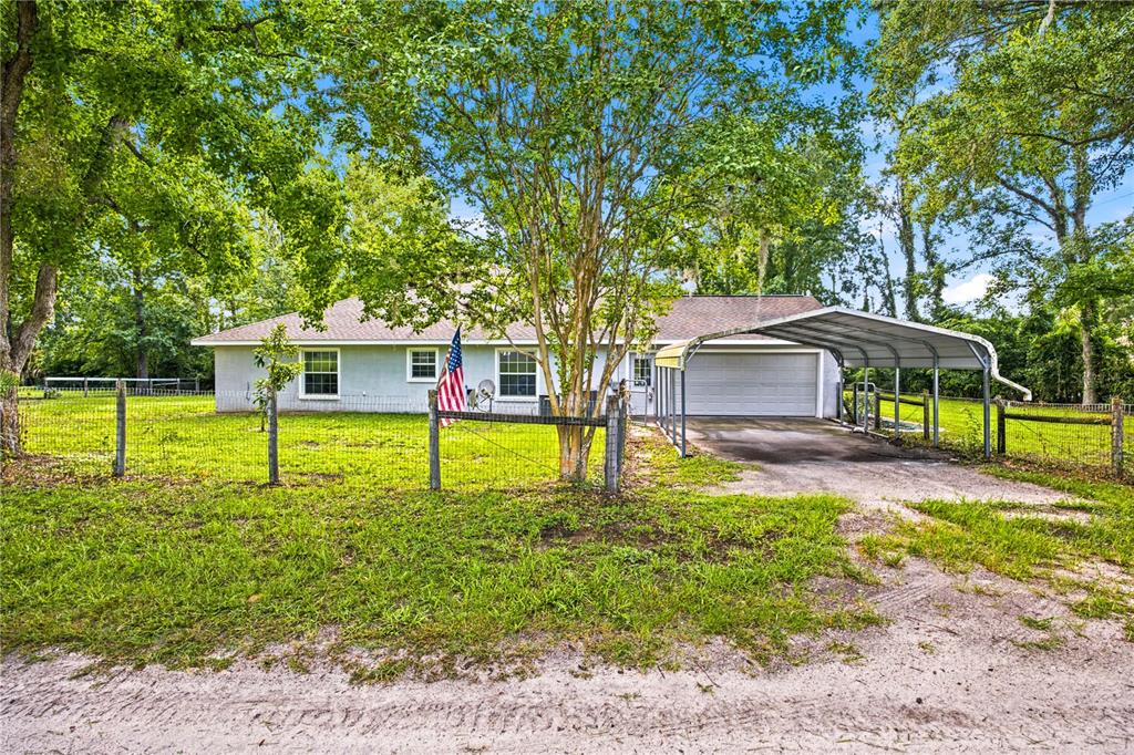 A single-story house with a metal carport, fenced yard, and American flag in a wooded area — 5259 SE 38TH STREET, OCALA, FL
