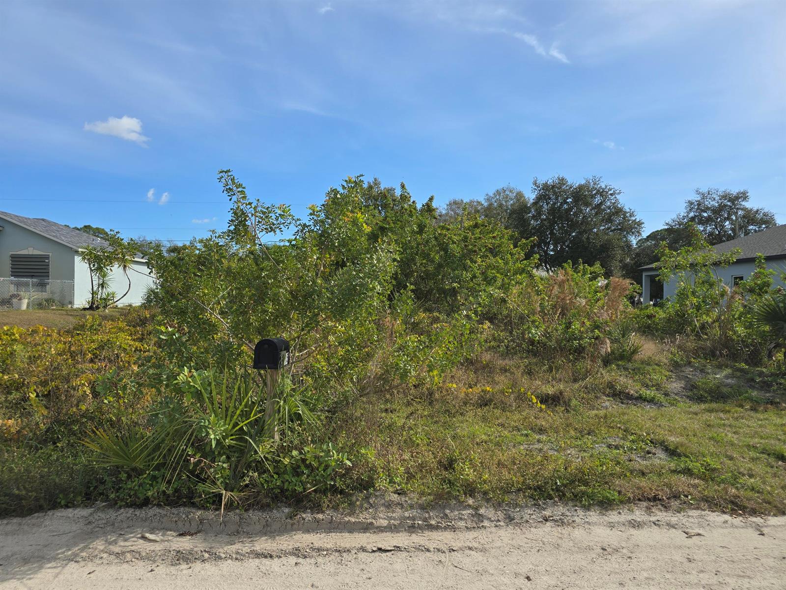 A vacant, overgrown residential lot with a mailbox stands between two houses under a blue sky — 8016 104th Court, Vero Beach FL 32967, VERO BEACH, FL