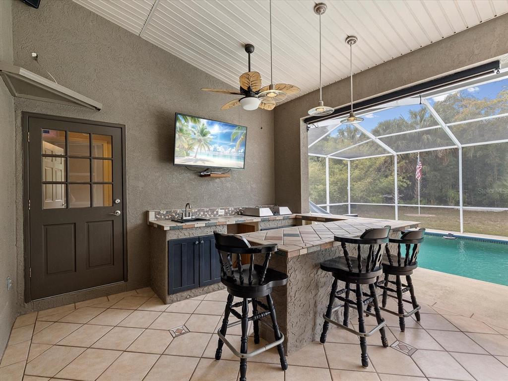 An outdoor kitchen with a tiled bar, three stools, and a television overlooks a pool — 8551 PRATT DRIVE, NEW PORT RICHEY, FL
