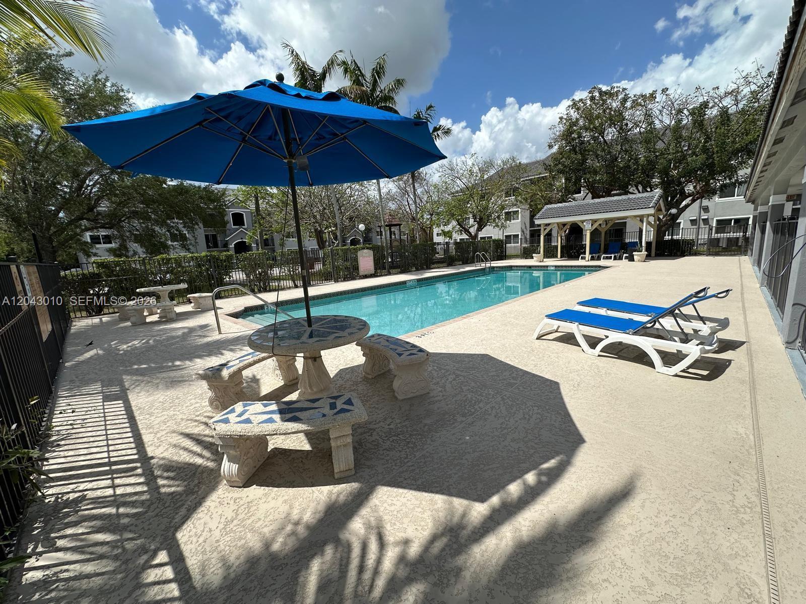 An outdoor swimming pool area features a blue umbrella, stone table, and lounge chairs — HOMESTEAD, FL