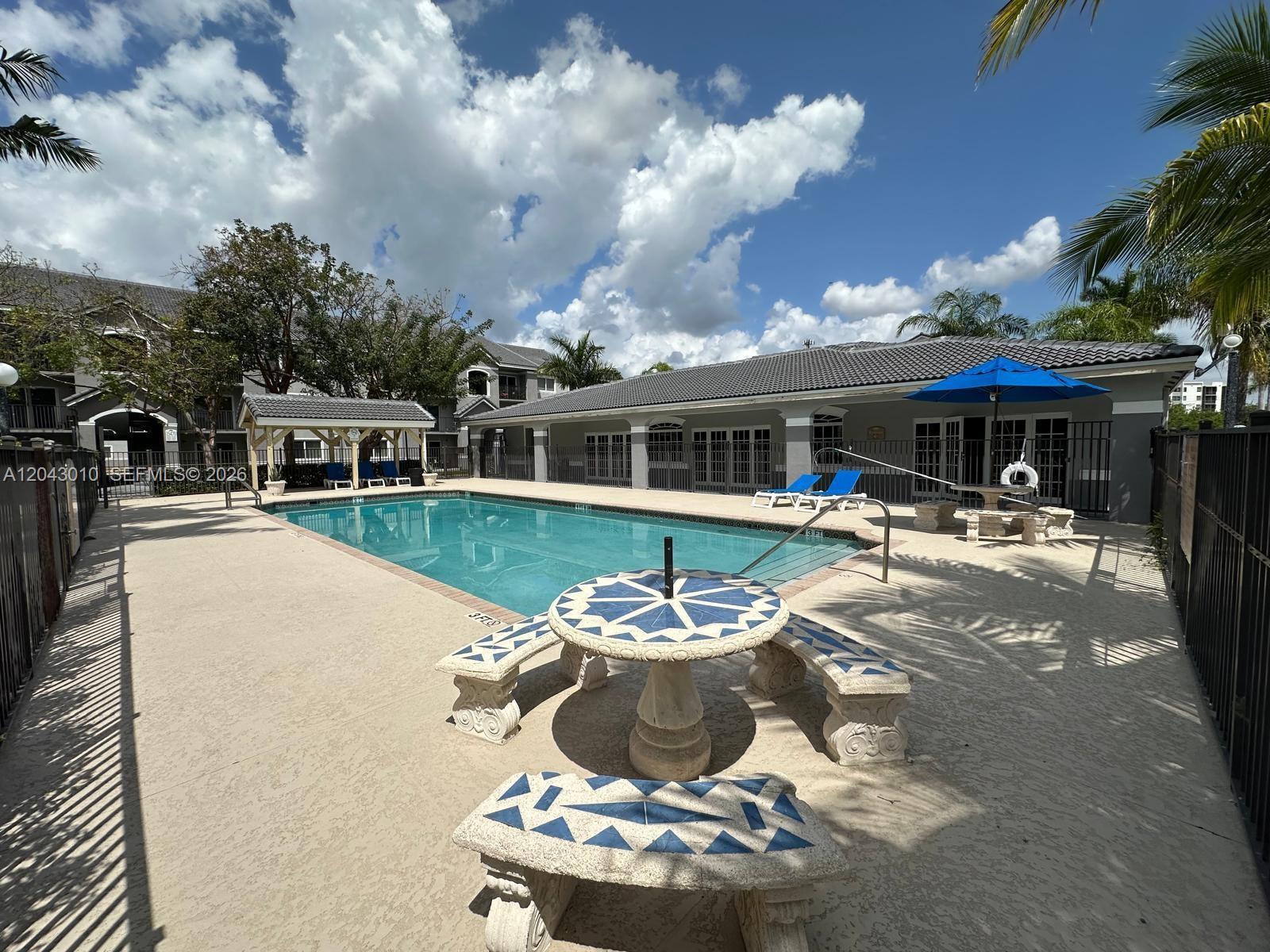 An outdoor swimming pool area features a tiled table with benches and a blue umbrella — HOMESTEAD, FL