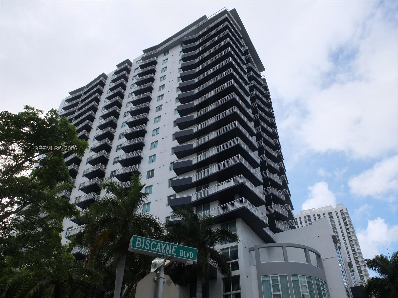 A low-angle view of a tall, modern apartment building with stacked balconies and palm trees — 275 NE 18th St # 1101, Miami FL 33132, MIAMI, FL