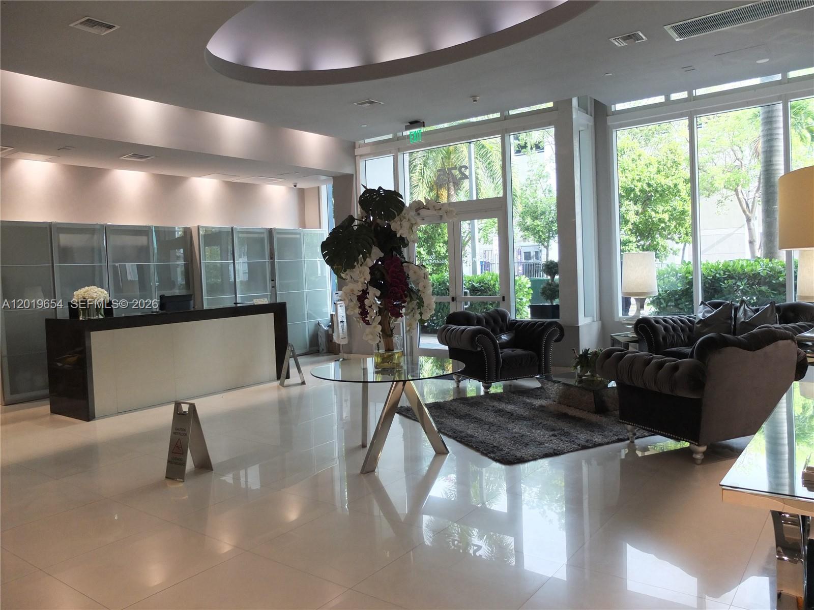 A modern lobby features a reception desk, plush seating, a glass table, and large windows — 275 NE 18th St # 1101, Miami FL 33132, MIAMI, FL