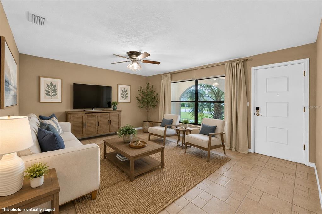 This virtually staged living room features tile flooring, a ceiling fan, and a front door — 475 WOODSTOCK DRIVE, PORT ORANGE, FL